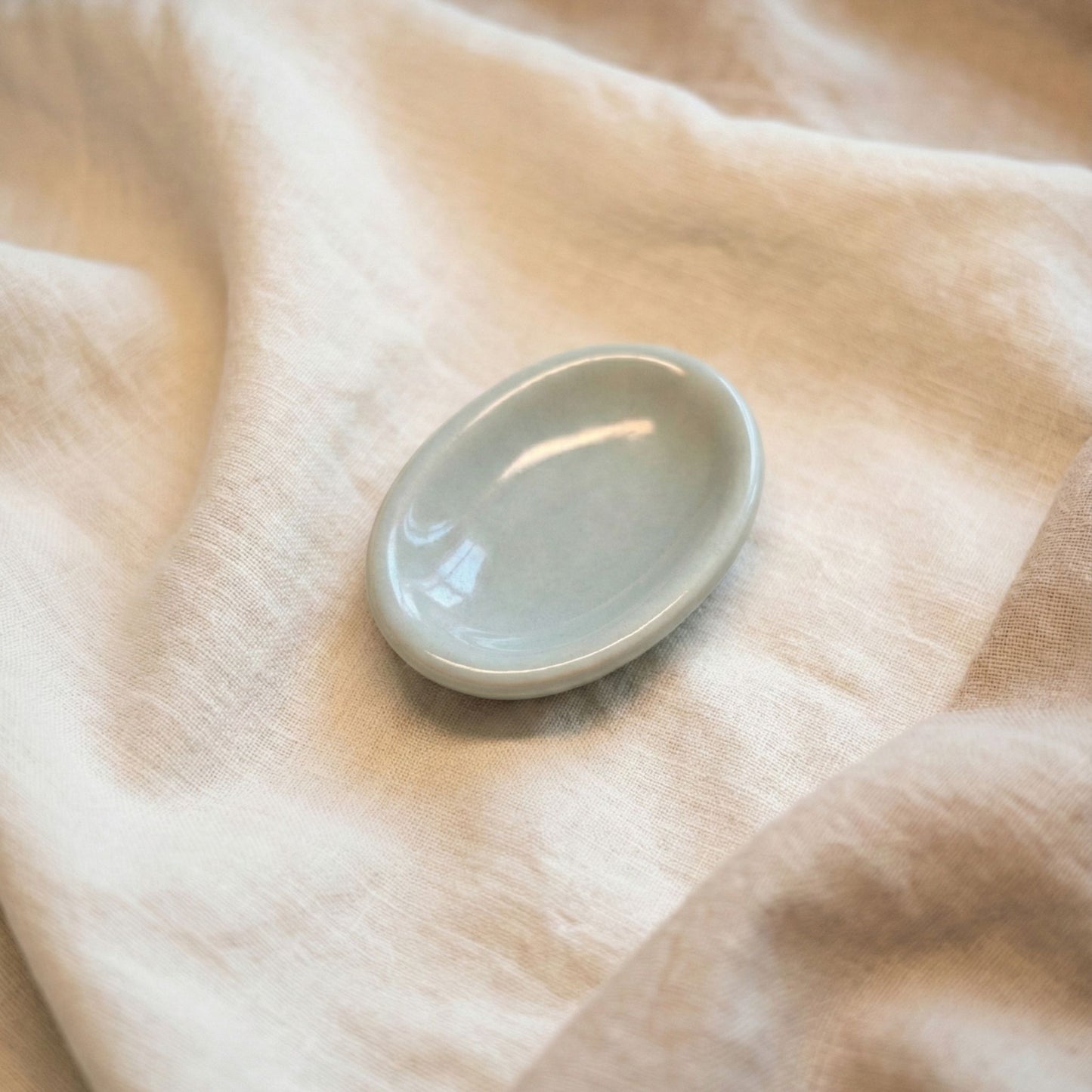 A minimal handmade ceramic worry stone in a soft grey glaze placed on a neutral fabric background, designed for grounding, anxiety relief, and calming tactile use.