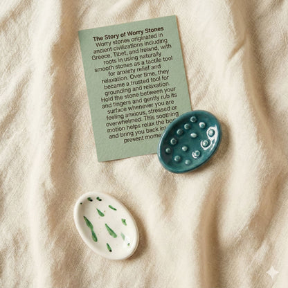 A set of handmade ceramic worry stones with tactile textures placed on soft fabric, alongside a green story card explaining the history and calming benefits of worry stones. Includes a teal textured stone and a white stone with green accents, designed for grounding and anxiety relief.