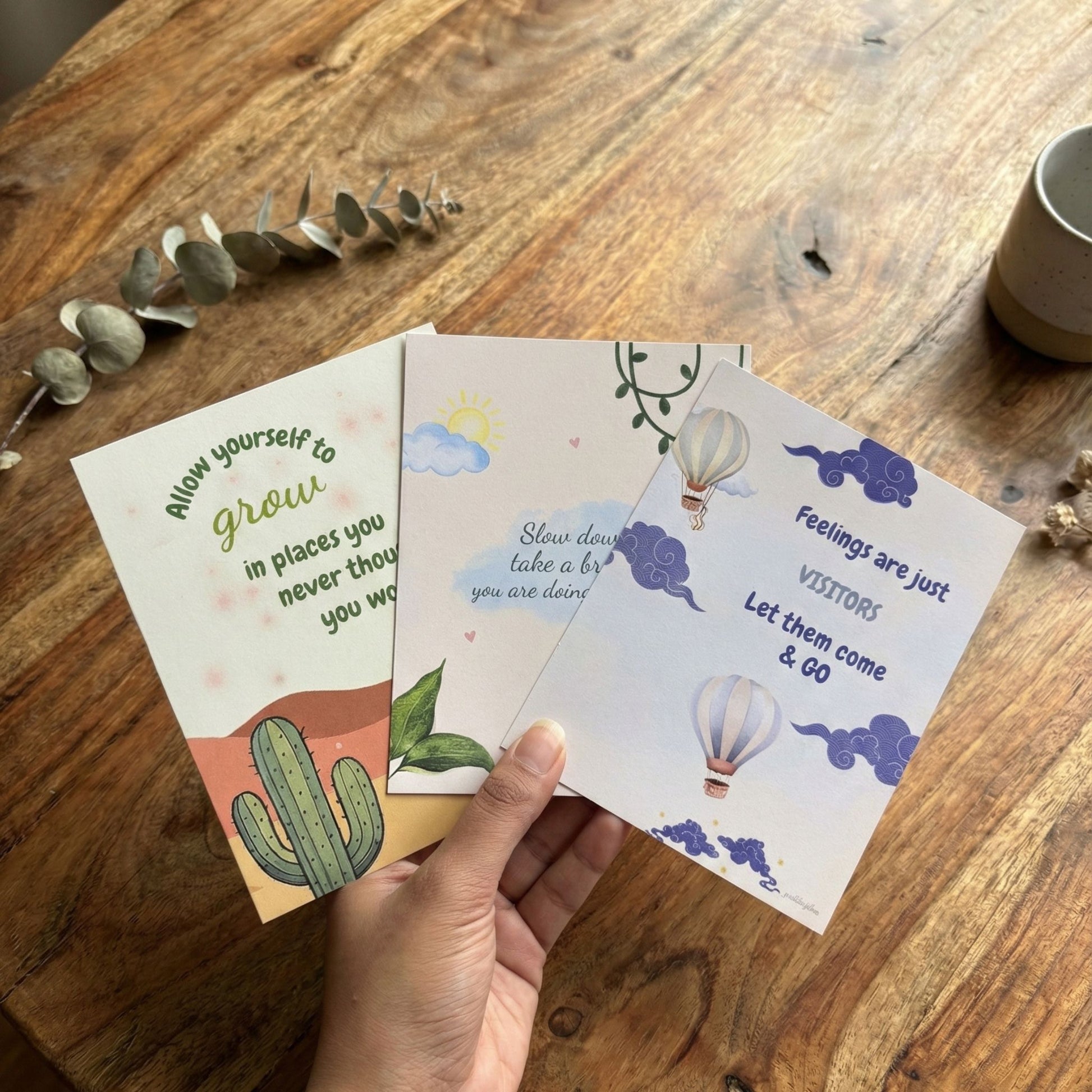 Set of three Guiding Koi positive affirmation postcards held over a wooden table, with calming illustrations and motivational quotes designed for mindfulness and emotional wellbeing.