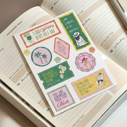 Positive affirmation and motivational sticker sheet placed on an open journal, featuring uplifting quotes like I Am Worthy, It’s Okay to Take It Slow, and Doing What You Love Is Never a Waste of Time.